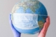 A globe of the world sporting a surgical mask held up by a pair of hands in purple and orange surgical gloves.